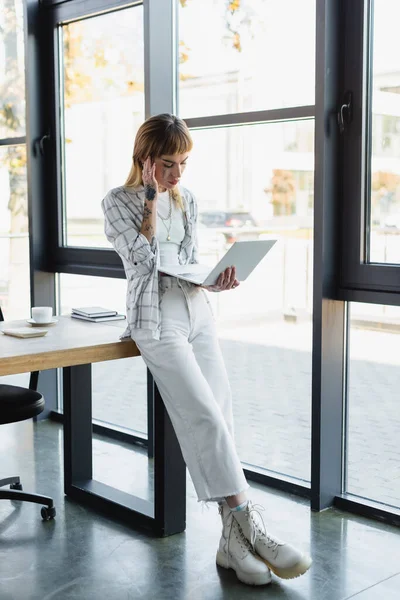 Vista completa de la mujer tatuada en ropa elegante usando el ordenador portátil mientras está de pie en el lugar de trabajo - foto de stock