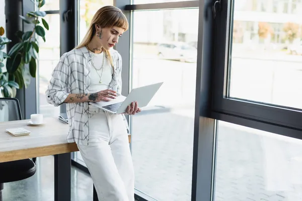 Young and stylish businesswoman with tattoo using laptop while standing at workplace in office — Stock Photo