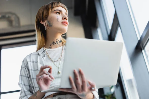 Vista basso angolo di donna tatuata con computer portatile offuscata guardando lontano mentre in piedi in ufficio — Foto stock