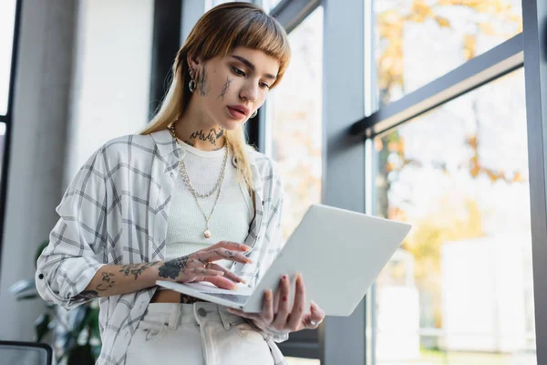 Stylish tattooed businesswoman standing in office and typing on laptop — Stock Photo