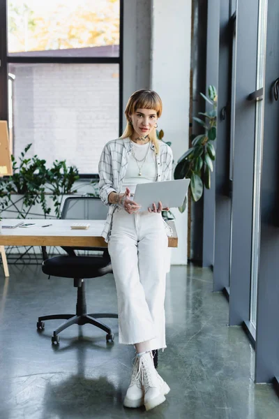 Vue complète de la femme d'affaires tatouée avec ordinateur portable debout sur le lieu de travail et regardant la caméra — Photo de stock