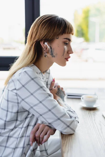 Vista lateral da mulher tatuada no fone de ouvido sem fio sentado no local de trabalho com os braços cruzados — Fotografia de Stock