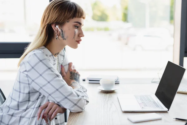 Joven mujer de negocios tatuada en auricular sentado con brazos cruzados cerca de la computadora portátil con pantalla en blanco - foto de stock