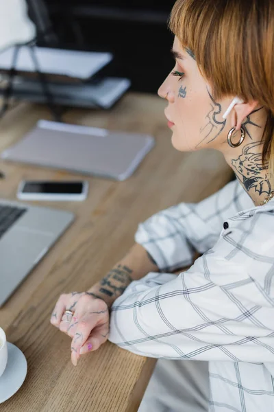 Giovane donna tatuata in auricolare wireless seduta sul posto di lavoro vicino a gadget sfocati — Foto stock