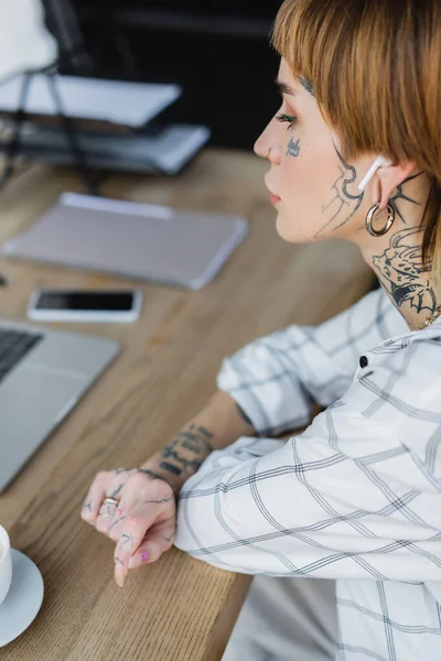 Junge tätowierte Frau mit drahtlosem Kopfhörer sitzt am Arbeitsplatz in der Nähe verschwommener Geräte — Stockfoto