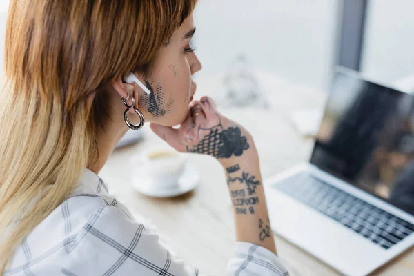 Femme d'affaires tatouée dans un écouteur sans fil regardant un ordinateur portable flou dans le bureau — Photo de stock