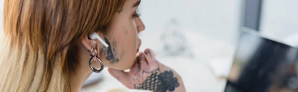 Joven mujer tatuada en auriculares inalámbricos mirando borrosa portátil en la oficina, pancarta - foto de stock