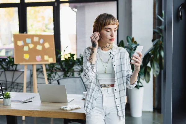 Trendy businesswoman with tattoo adjusting earphone during video call on cellphone — Stock Photo