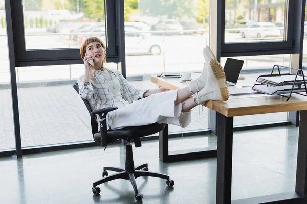 Elegante mujer de negocios tatuada sentada con piernas en el escritorio mientras habla en el teléfono inteligente en la oficina - foto de stock
