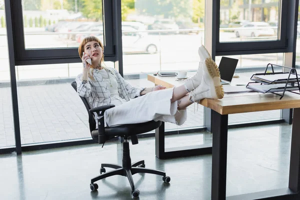 Élégant tatoué femme d'affaires assis avec les jambes sur le bureau tout en parlant sur smartphone dans le bureau — Photo de stock