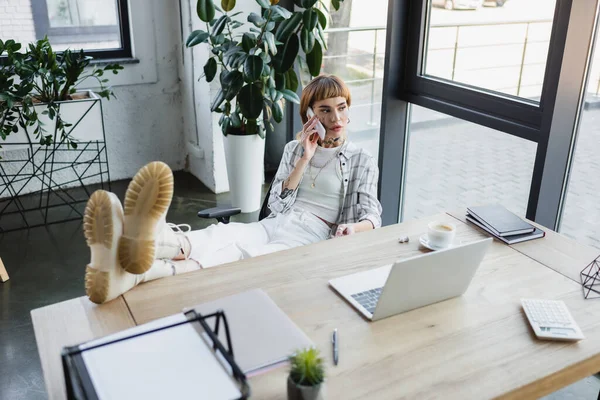 High angle view of tattooed woman with legs on desk talking on mobile phone in office — Stock Photo
