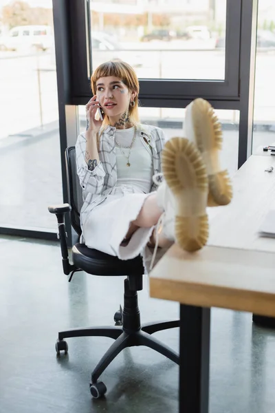 Elegante tatuado mulher sentado com pernas na mesa enquanto falando no celular no escritório — Fotografia de Stock