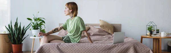 Joven mujer tatuada sentada en la cama cerca de la computadora portátil en el dormitorio moderno con plantas en maceta, pancarta - foto de stock