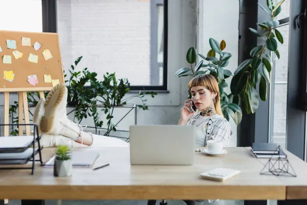 Trendy businesswoman calling on cellphone while sitting with legs on desk near blurred laptop — Stock Photo