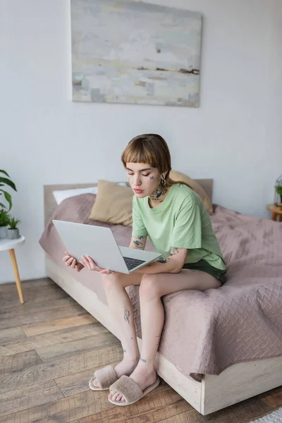 Mujer bonita con cuerpo tatuado trabajando en el ordenador portátil mientras está sentado en la cama en casa - foto de stock