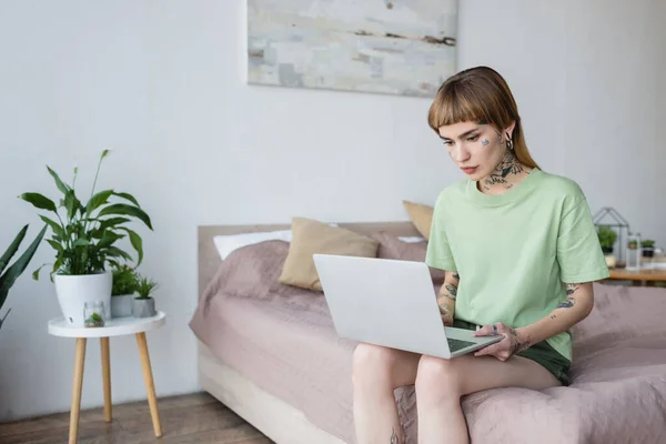 Jeune femme avec tatouage à l'aide d'un ordinateur portable tout en étant assis sur le lit à la maison — Photo de stock