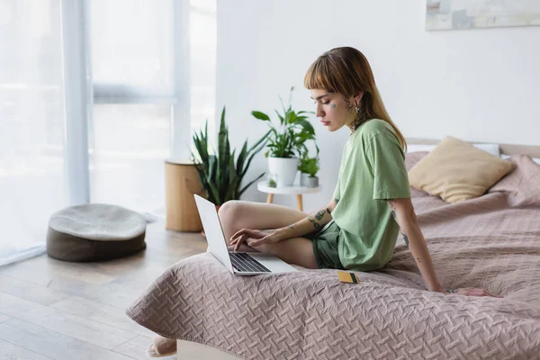 Tätowierte Frau sitzt in der Nähe der Kreditkarte auf dem Bett und benutzt Laptop — Stockfoto
