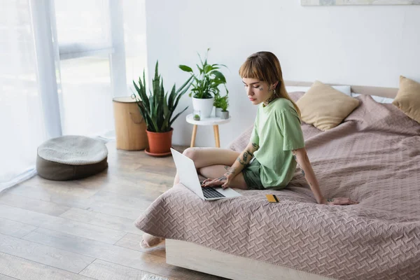 Frau mit tätowiertem Körper tippt auf Laptop, während sie in der Nähe der Kreditkarte auf dem Bett sitzt — Stockfoto