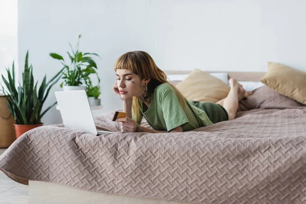 Татуйована жінка лежить на ліжку з ноутбуком і кредитною карткою біля розмитих рослин у спальні — стокове фото