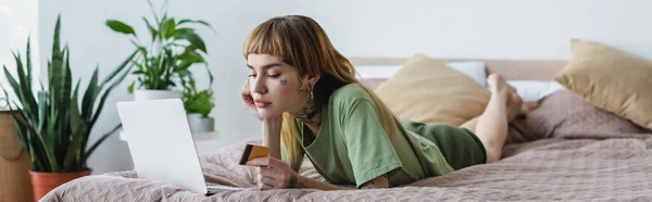 Joven mujer tatuada sosteniendo tarjeta de crédito mientras está acostado en la cama cerca de la computadora portátil, pancarta — Stock Photo