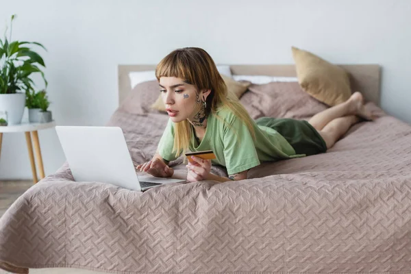 Mujer joven con el cuerpo tatuado acostado en la cama con tarjeta de crédito y portátil — Stock Photo