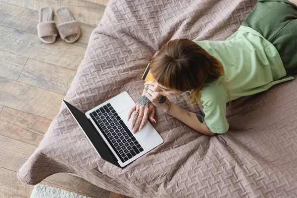 Vista aérea da mulher tatuada usando laptop enquanto deitada na cama com cartão de crédito — Fotografia de Stock