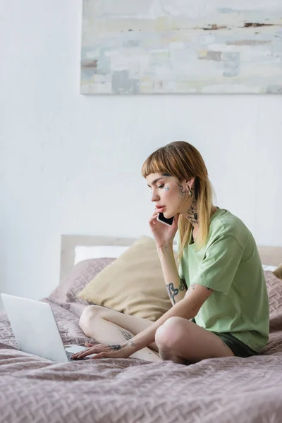 Tatuado mulher falando no smartphone enquanto usando laptop na cama em casa — Fotografia de Stock