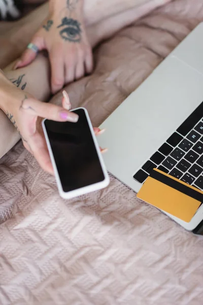 Vista parcial de la mujer borrosa con el teléfono inteligente de la celebración del tatuaje con la pantalla en blanco cerca de la computadora y la tarjeta de crédito - foto de stock