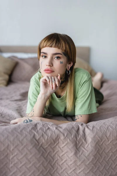 Mujer con tatuaje y piercing descansando en la cama en casa y mirando a la cámara - foto de stock