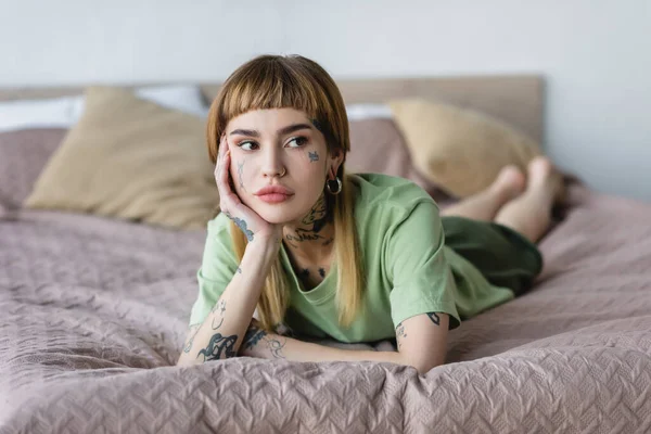 Junge Frau mit Tätowierung und Piercing schaut weg, während sie sich im Schlafzimmer ausruht — Stockfoto