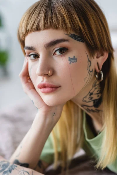 Vista de cerca de la mujer bonita con tatuaje y piercing mirando a la cámara en casa - foto de stock