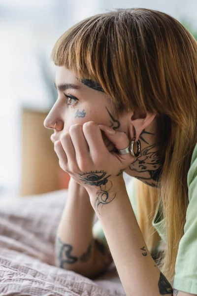 Vista lateral de mujer tatuada con piercing mirando hacia otro lado en la cama en casa - foto de stock