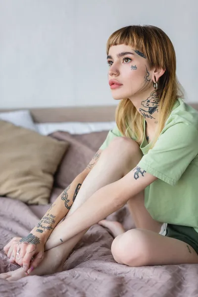 Joven mujer tatuada mirando hacia otro lado mientras está sentada en la cama en casa - foto de stock