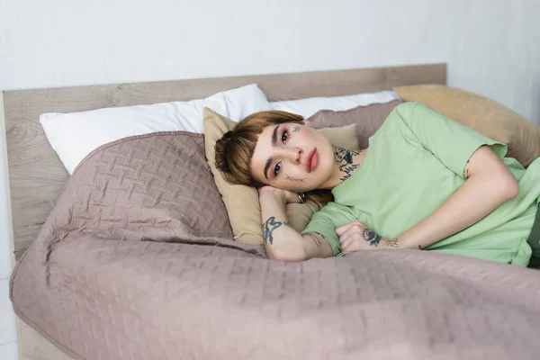 Hübsche tätowierte Frau blickt in die Kamera, während sie im Schlafzimmer auf Kissen liegt — Stockfoto