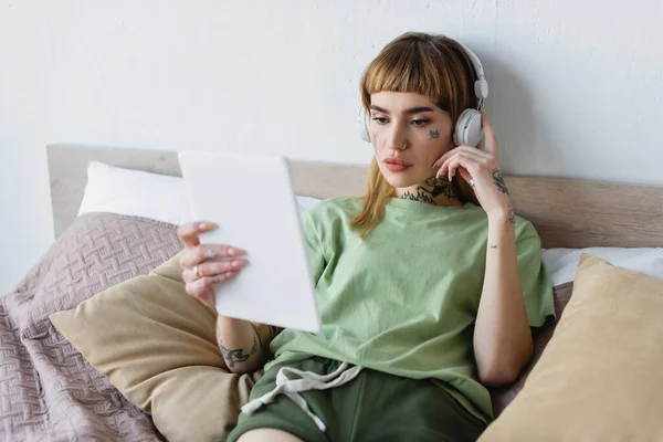 Young tattooed woman in headphones looking at blurred digital tablet while listening podcast in bedroom — Stock Photo