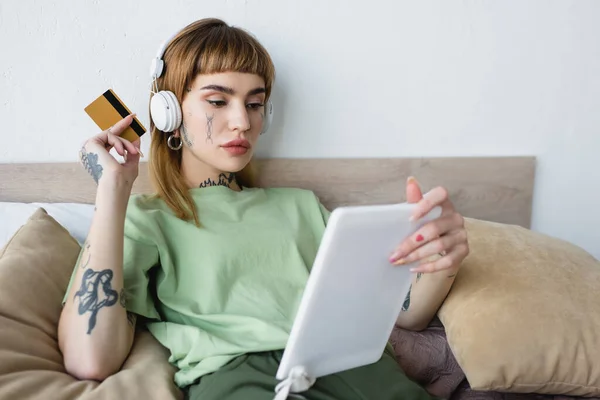 Mujer tatuada en auriculares con tarjeta de crédito mientras usa la tableta digital en el dormitorio - foto de stock