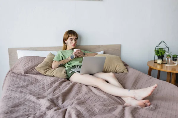 Full length view of young tattooed woman drinking tea and watching film on laptop in bed — Fotografia de Stock