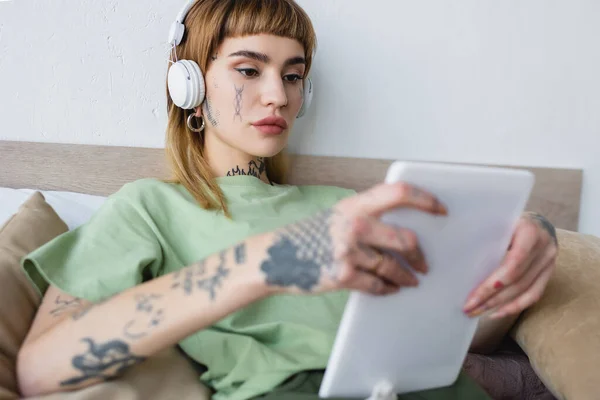 Молода татуйована жінка з розмитим цифровим планшетом прослуховування подкасту в спальні — стокове фото