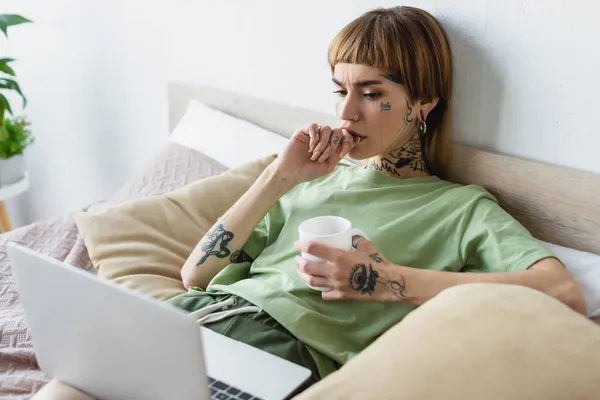 Joven mujer preocupada con el cuerpo tatuado viendo la película en el ordenador portátil borroso - foto de stock
