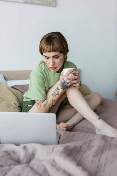 Concentrated woman with tattoo holding cup of tea while watching movie on blurred laptop on bed — Stock Photo