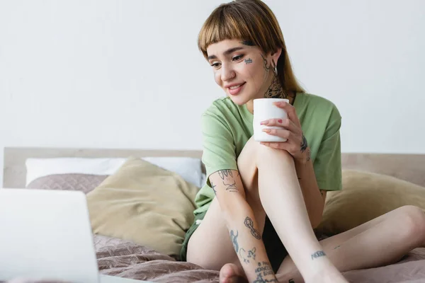 Femme heureuse avec corps tatoué tenant tasse de thé tout en regardant un film sur ordinateur portable flou — Photo de stock