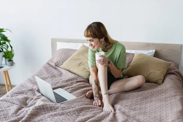 Young tattooed woman sitting on bed and watching movie on laptop — Stock Photo