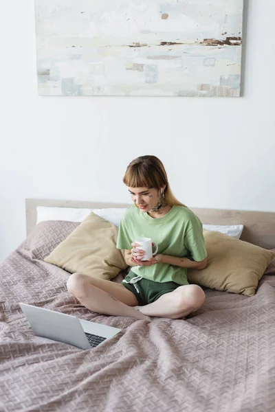 Tattooed woman sitting on bed with crossed legs and watching film on laptop — Stock Photo