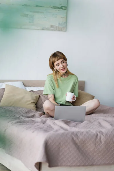 Sorrindo mulher tatuada sentada na cama com xícara de chá e assistindo filme no computador — Fotografia de Stock