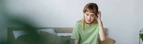 Tattooed woman touching head while sitting in bedroom on blurred foreground, banner — Stock Photo