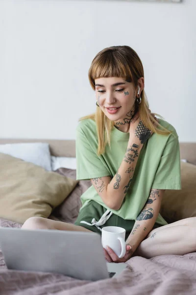 Положительная женщина с татуированным телом смотрит фильм на ноутбуке в спальне — стоковое фото
