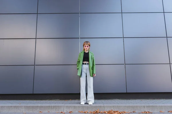 Ganzkörperansicht der tätowierten Frau in stilvoller Herbstkleidung und Brille in der Nähe der grauen Mauer auf der Stadtstraße — Stockfoto