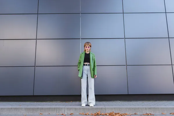 Full length view of tattooed woman in stylish autumn clothes and eyeglasses near grey wall on city street — Stock Photo