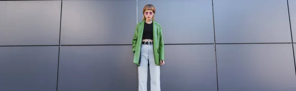 Vorderseite tätowierte Frau in grüner Lederjacke, die neben grauer Wand steht, Banner — Stockfoto