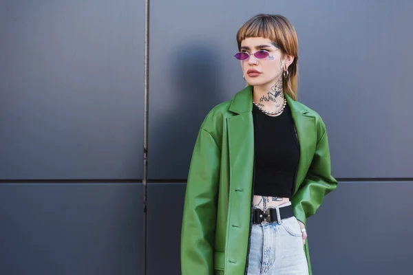 Tattooed woman in green jacket standing with hand on hip and looking away near grey wall — Stock Photo